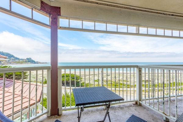 A balcony view showing a table and chairs overlooking a sandy beach and ocean under a partly cloudy sky.