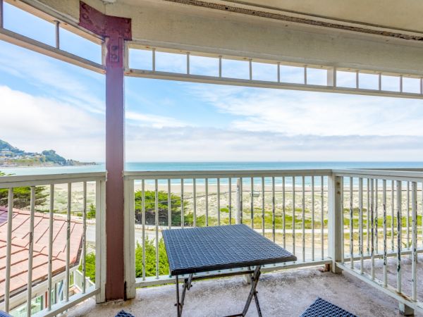 A balcony view showing a table and chairs overlooking a sandy beach and ocean under a partly cloudy sky.