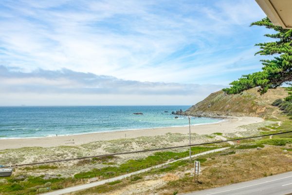 A scenic view of a beach with gentle waves, a sandy shore, a hillside, and some greenery under a partly cloudy sky.