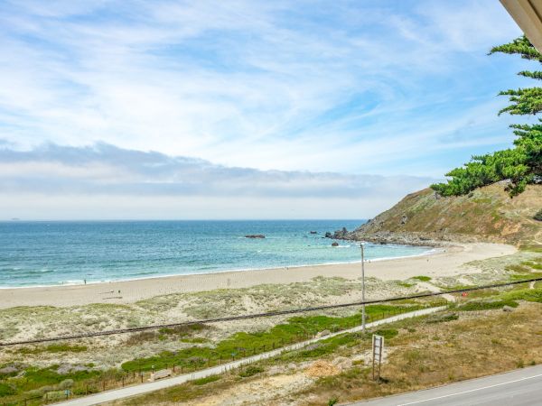 A scenic view of a beach with gentle waves, a sandy shore, a hillside, and some greenery under a partly cloudy sky.