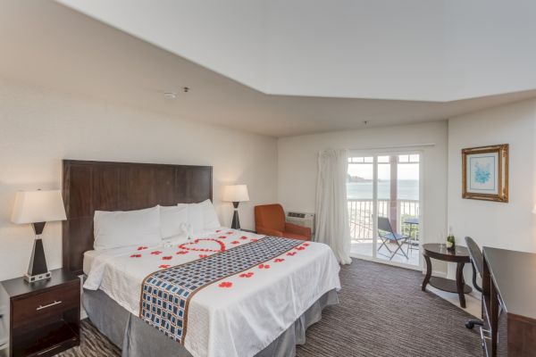 A hotel room with a king-sized bed decorated with rose petals, bedside lamps, a chair, a desk, and a balcony with ocean view.