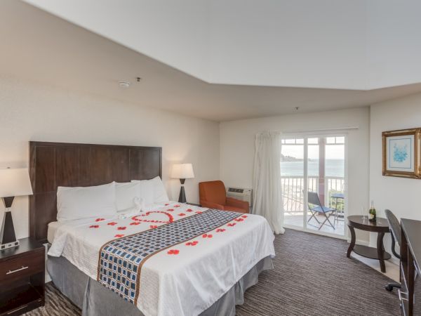 A hotel room with a king-sized bed decorated with rose petals, bedside lamps, a chair, a desk, and a balcony with ocean view.
