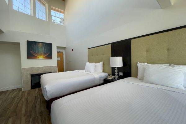 This image shows a hotel room with two beds, a lamp, a painting on the wall, and a high ceiling with large windows and wood flooring.