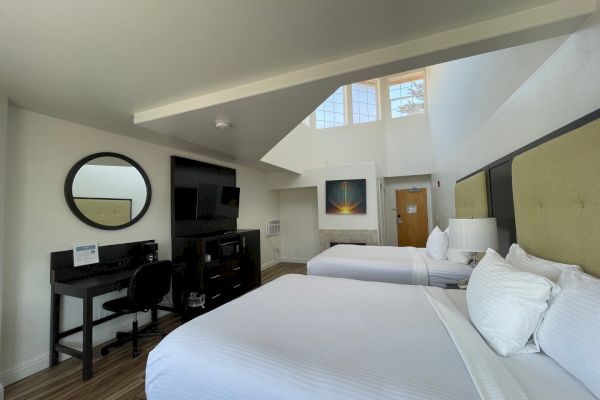 A modern hotel room with two beds, a desk, TV, and wall art. High ceilings and large windows enhance the space's brightness.