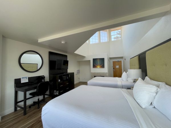 A modern hotel room with two beds, a desk, TV, and wall art. High ceilings and large windows enhance the space's brightness.