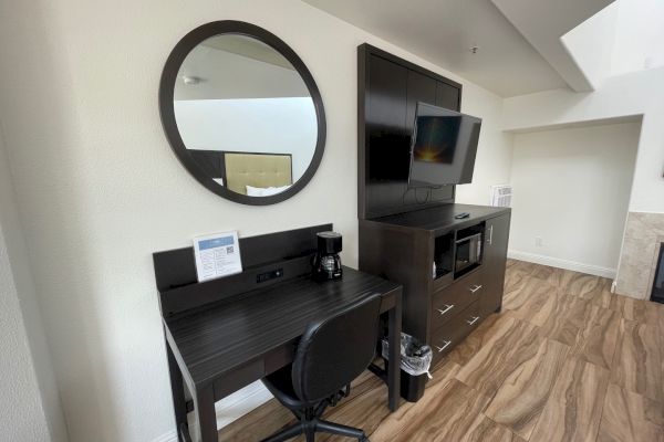 A modern hotel room with a desk, chair, round mirror, TV, microwave, and wooden flooring.