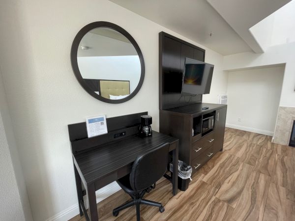 A modern hotel room with a desk, chair, round mirror, TV, microwave, and wooden flooring.