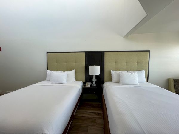 The image shows a hotel room with two neatly made beds, a lamp, and a small bedside table between them.
