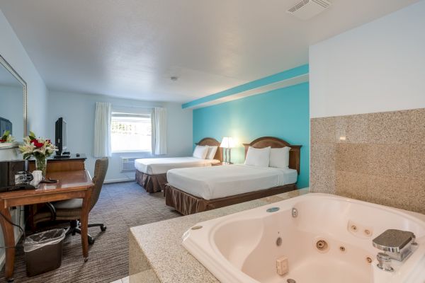 A hotel room with two beds, a desk, a TV, and an in-room jacuzzi. The decor is simple with a blue accent wall and warm lighting.