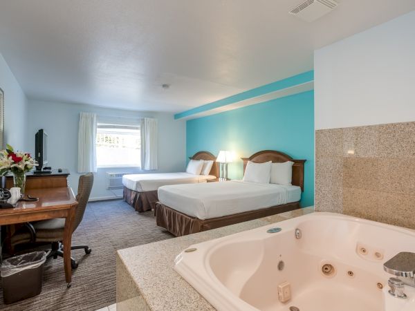 A hotel room with two beds, a desk, a TV, and an in-room jacuzzi. The decor is simple with a blue accent wall and warm lighting.