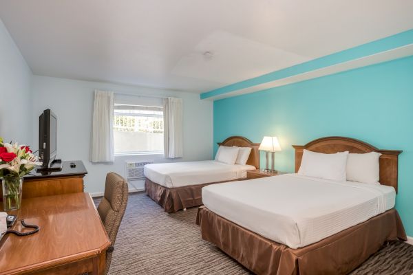 A hotel room with two beds, a TV, a desk with a chair, and a window. The walls are light blue, and there's a lamp between the beds.