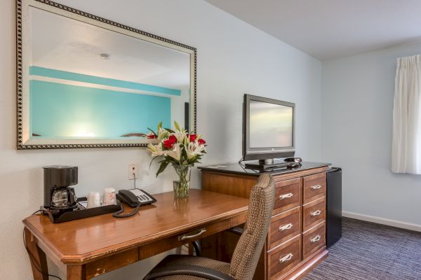 A hotel room desk with a mirror, flowers, coffee maker, phone, chair, and a TV on a dresser.