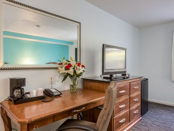 A hotel room desk with a mirror, flowers, coffee maker, phone, chair, and a TV on a dresser.