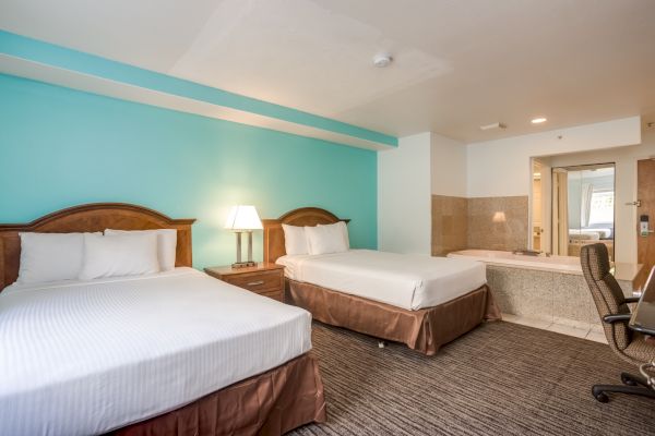 The image shows a hotel room with two beds, a nightstand with a lamp, a chair, and a large bathtub in the corner near the bathroom.