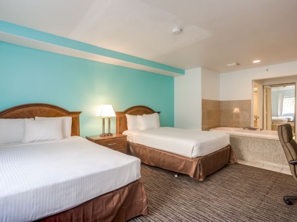 The image shows a hotel room with two beds, a nightstand with a lamp, a chair, and a large bathtub in the corner near the bathroom.