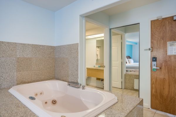 The image shows a hotel room with a large bathtub, a door, and a view into another room with a bed and vanity.