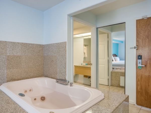 The image shows a hotel room with a large bathtub, a door, and a view into another room with a bed and vanity.