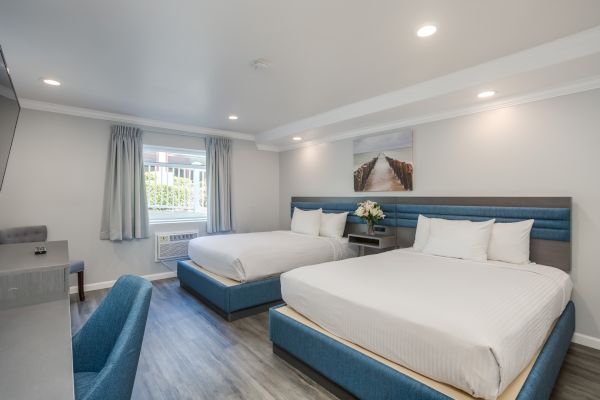 A modern hotel room with two large beds, blue accents, desk, chair, wall art, and window with curtains. Bright lighting, tidy ambiance.