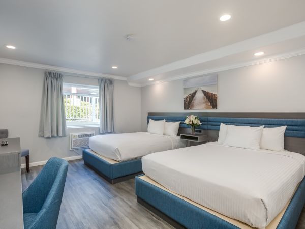 A modern hotel room with two large beds, blue accents, desk, chair, wall art, and window with curtains. Bright lighting, tidy ambiance.