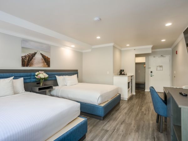 The image shows a hotel room with two double beds, a desk, a chair, wall art, and a small kitchenette area.