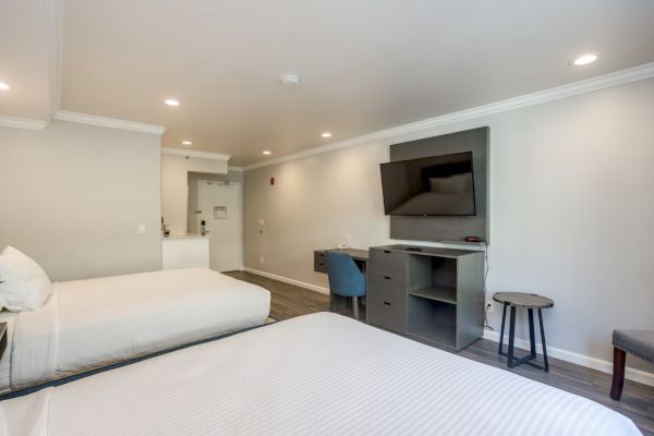 The image shows a hotel room with two beds, a TV on the wall, a desk, and a chair in a modern style.