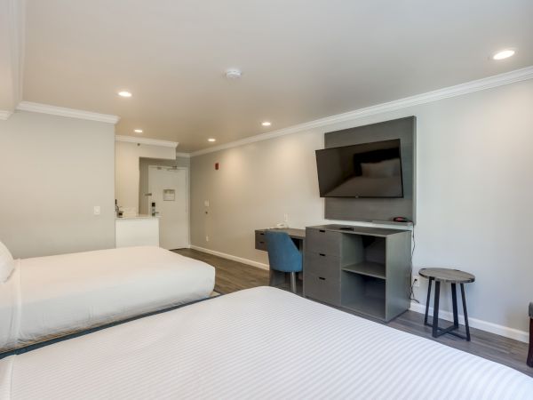 The image shows a hotel room with two beds, a TV on the wall, a desk, and a chair in a modern style.