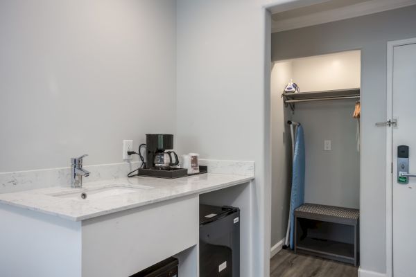 The image shows a hotel room corner with a kitchenette area, mini fridge, coffee maker, and a closet space with hangers and an ironing board.