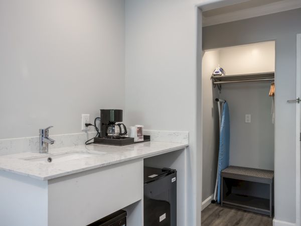 The image shows a hotel room corner with a kitchenette area, mini fridge, coffee maker, and a closet space with hangers and an ironing board.