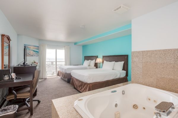 A hotel room with two beds, a TV, desk, and a jacuzzi. The room has a bright, beachy feel with a view through the sliding doors.