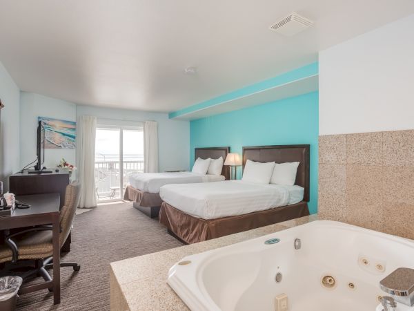 A hotel room with two beds, a TV, desk, and a jacuzzi. The room has a bright, beachy feel with a view through the sliding doors.