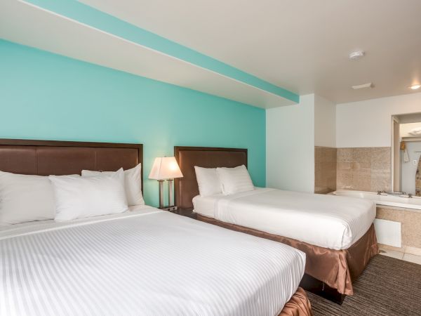 The image shows a hotel room with two beds, a nightstand with a lamp, and a whirlpool tub in the background.