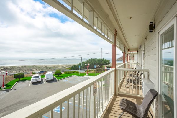 A balcony with chairs overlooks a parking lot, green lawn, and beach. The ocean and sky create a scenic background view from the building.