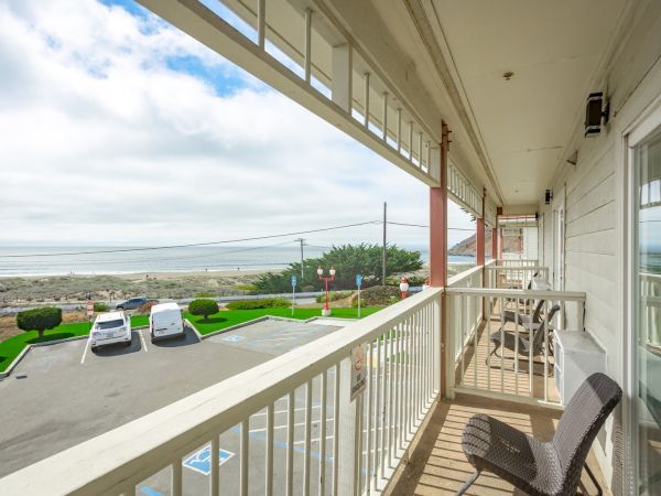 A balcony with chairs overlooks a parking lot, green lawn, and beach. The ocean and sky create a scenic background view from the building.