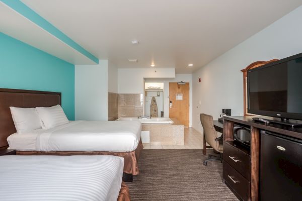 The image shows a hotel room with two beds, a TV, dresser, desk, chair, and a bathroom with a bathtub at the back.