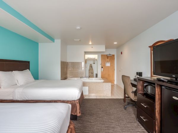 The image shows a hotel room with two beds, a TV, dresser, desk, chair, and a bathroom with a bathtub at the back.