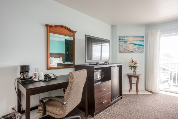 The image shows a hotel room with a desk, chair, mirror, TV stand, microwave, TV, artwork, a small table with flowers, and a sliding glass door.