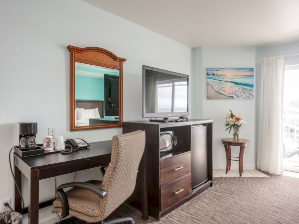 The image shows a hotel room with a desk, chair, mirror, TV stand, microwave, TV, artwork, a small table with flowers, and a sliding glass door.