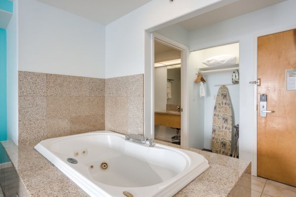 The image shows a bathroom with a large bathtub, an ironing board, and a closet space. There's a wooden door and tiled surfaces.