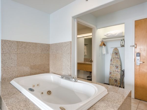 The image shows a bathroom with a large bathtub, an ironing board, and a closet space. There's a wooden door and tiled surfaces.