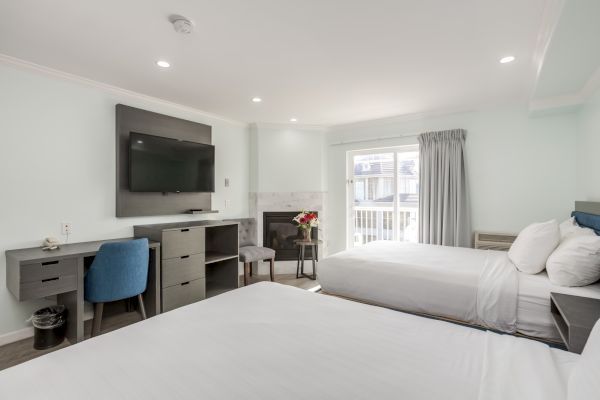 A modern hotel room with two beds, a flat-screen TV, desk, chair, fireplace, and flowers on a table by a window.