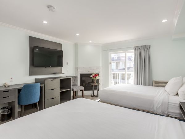 A modern hotel room with two beds, a flat-screen TV, desk, chair, fireplace, and flowers on a table by a window.
