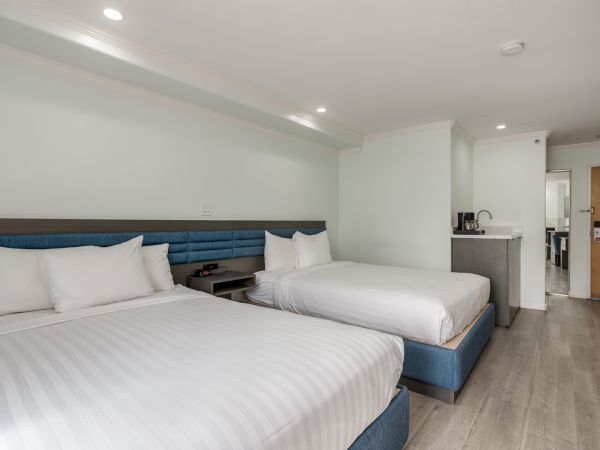 The image shows a hotel room with two neatly made double beds, modern decor, wooden flooring, and a small kitchenette area in the corner.