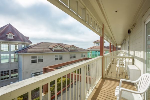 A balcony with white railings and chairs overlooks neighboring buildings with tiled roofs under a partly cloudy sky.