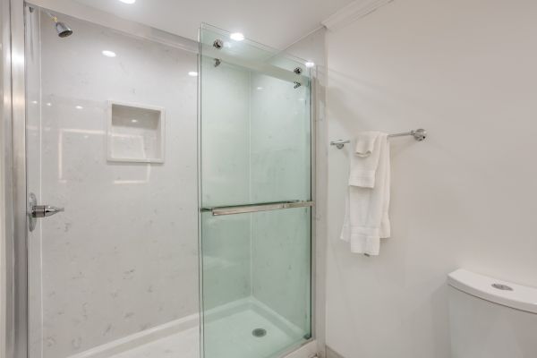 A modern bathroom with a glass shower enclosure, white walls, towels on a rack, and a toilet visible on the right side of the image.