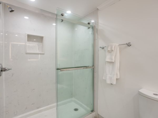 A modern bathroom with a glass shower enclosure, white walls, towels on a rack, and a toilet visible on the right side of the image.