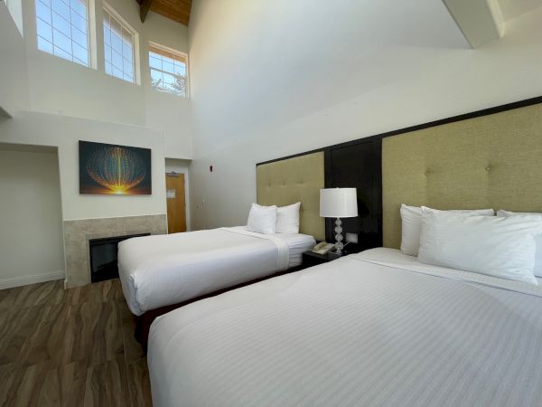 The image shows a hotel room with two neatly made beds, a lamp between them, a painting on the wall, and large windows.