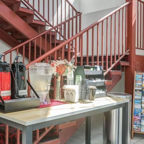 A refreshment station with coffee dispensers and brochures on a rack is set up near a red staircase.