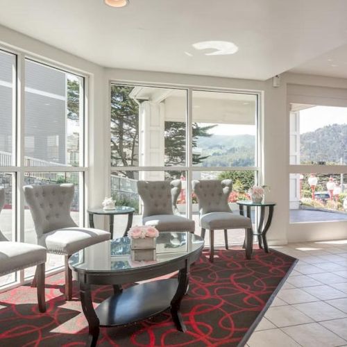 A bright lobby area with chairs and glass tables on a patterned rug. Large windows offer views of the outside and parked cars.