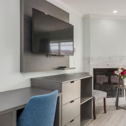 The image shows a modern room with a wall-mounted TV, desk, blue chair, fireplace, side table with flowers, and a sliding glass door.