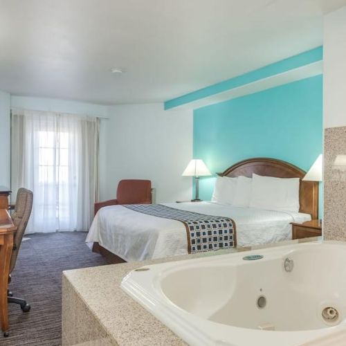 A hotel room featuring a bed, desk, chair, TV, and a hot tub near the entrance. Bright lighting and a vibrant blue accent wall stand out.